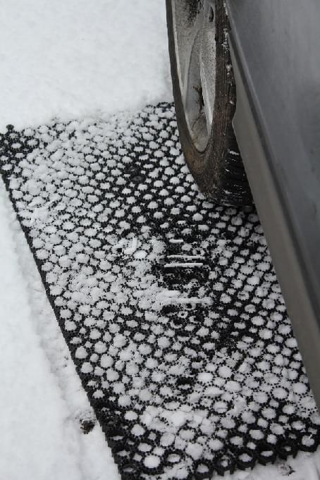 Zdjęcie przedstawia fragment koła zaparkowany na śniegowej macie antypoślizgowej, leżącej na śniegu.